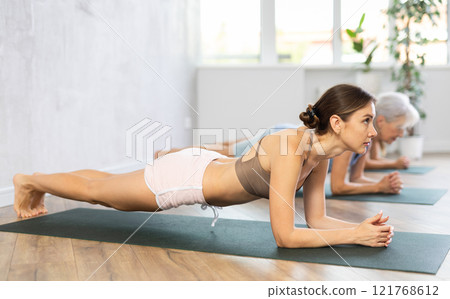 Three generations of women perform elbow plank Sampatasana in gym 121768612