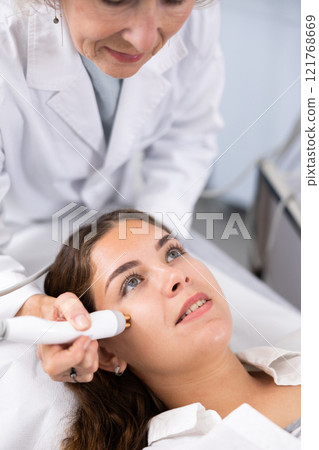 Elderly beautician doctor doing RF lifting procedure for flawless woman face laying in beauty salon. Hardware cosmetology. Elderly beautician doctor doing RF lifting procedure for flawless woman face laying in beauty salon. Hardware cosmetology. 121768669