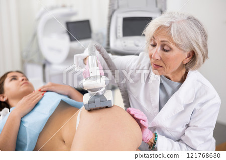 Old woman specialist doing LPG massage of hips of young woman Old woman specialist doing LPG massage of hips of young woman 121768680