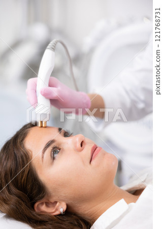 Unrecognizable beautician doctor doing RF lifting procedure for flawless woman face laying in beauty salon. Hardware cosmetology 121768731