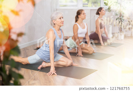 Elderly woman stretching in seated asana Ardha Kapotasana in yoga studio 121768755