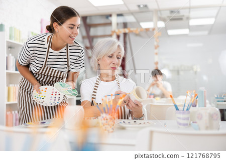 Young girl assisting senior woman painting ceramic mug in pottery class 121768795