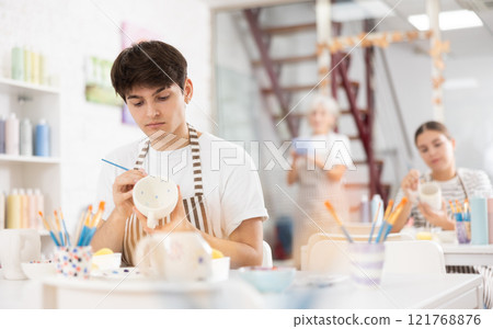 Focused guy enthusiastically painting on ceramic cup in pottery class 121768876