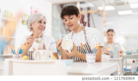 Cheerful elderly woman and guy painting ceramic mugs during pottery masterclass 121768904