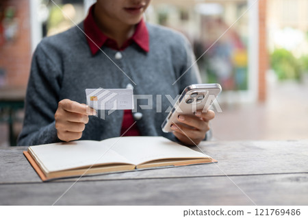 A cropped image of a woman holding a smartphone and a credit card while sitting at an outdoor table. A cropped image of a woman holding a smartphone and a credit card while sitting at an outdoor table. 121769486