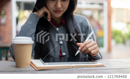 An Asian woman holding a stylus pen, using a digital tablet while sitting at an outdoor table. 121769488