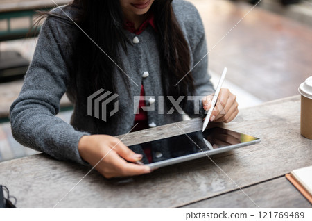An Asian woman holding a stylus pen, using a digital tablet while sitting at an outdoor table. 121769489