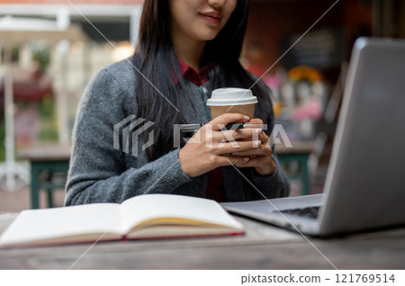 A female student studying online on her laptop while sitting at an outdoor cafe table. 121769514