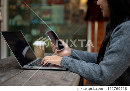 A woman holding her smartphone while working on her laptop, sitting at a table outdoors. 121769518
