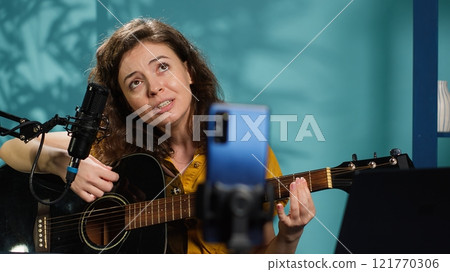 Talented woman playing music on guitar during livestream with audience online. Musician using musical instrument, having fun doing concert for fans producing entertaining songs, camera A Talented woman playing music on guitar during livestream with audience online. Musician using musical instrument, having fun doing concert for fans producing entertaining songs, camera A 121770306