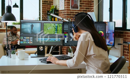 Asian filmmaker woman putting headphones on and working on video production at a home office, mixing video clips. Office setup with a software perfect for color grading in post production. Camera A. 121770311
