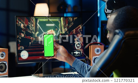 Man texting friends on green screen phone while playing first person shooter videogame at home. Gamer looking at mockup smartphone display, enjoying online discussion with mates 121770321