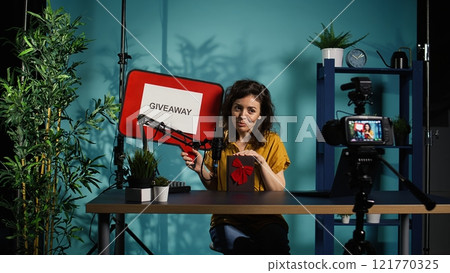 Media star holding empty copy space sign doing giveaway, offering packaged present. Woman does influencer marketing by offering prizes during internet contest, filming with professional gear, camera B Media star holding empty copy space sign doing giveaway, offering packaged present. Woman does influencer marketing by offering prizes during internet contest, filming with professional gear, camera B 121770325