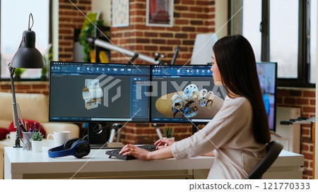 Professional industrial designer working with CAD software on dual monitors, creating 3D prototypes for a tech driven environment. 3D modeling software for innovative design. Camera B. 121770333