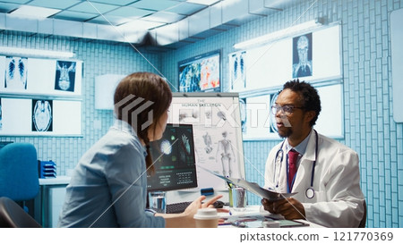 Female patient listening to a diagnostic report from doctor based on MRI scan results and x ray imaging, pointing at computer in medical cabinet. African american expert gives assistance. Camera B. Female patient listening to a diagnostic report from doctor based on MRI scan results and x ray imaging, pointing at computer in medical cabinet. African american expert gives assistance. Camera B. 121770369