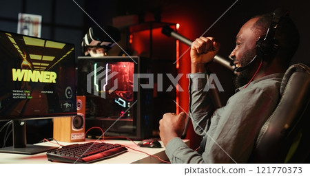 Man feeling happy while playing videogames on computer desktop with headphones on, celebrating victory. Cheerful gamer smiling after winning game on gaming PC while at home, camera B 121770373