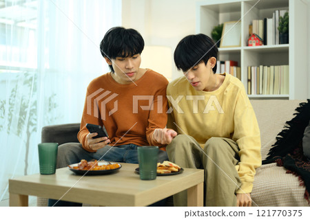 Cute gay couple chatting and enjoying snacks sitting sofa in bright living room 121770375