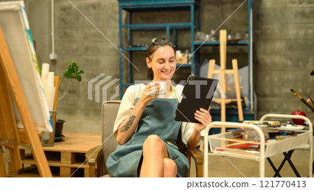Smiling female artist wearing apron enjoying a cup of coffee and using digital tablet in a creative studio Smiling female artist wearing apron enjoying a cup of coffee and using digital tablet in a creative studio 121770413