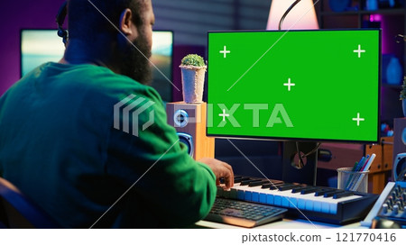 Musician operates on midi controller piano keys next to mockup screen, producing new songs with musical instruments and audio gear in home studio. Artist works with green screen. Camera A. 121770416