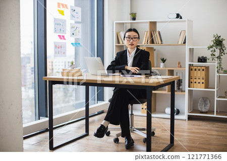 Asian businesswoman sitting confidently at office desk, wearing suit, glasses modern office setting with books, laptop, and decor, suggesting professional work environment. 121771366