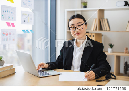 Young Asian businesswoman sitting at desk with laptop, taking notes. Professional setting includes bookshelves, diagrams on window. Elegant and focused workplace atmosphere. 121771368