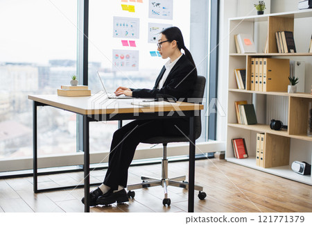 Asian businesswoman in professional attire working at laptop in modern office environment. Young adult focused on tasks, highlighting workplace productivity and focus. 121771379
