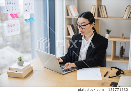 Asian businesswoman in professional attire working on laptop in bright office. Modern environment with bookshelves and paperwork. Concentrated expression reflecting focus and productivity. Asian businesswoman in professional attire working on laptop in bright office. Modern environment with bookshelves and paperwork. Concentrated expression reflecting focus and productivity. 121771380