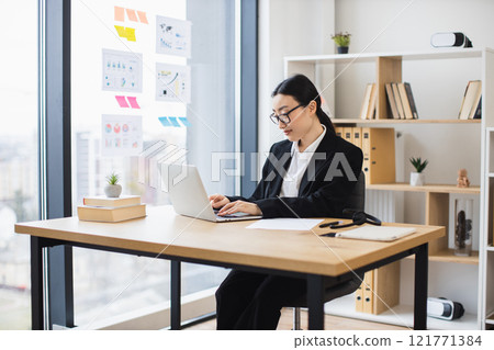 Asian businesswoman wearing glasses working on laptop at modern office desk. Professional atmosphere with natural light and organized workspace. Focused and productive environment. Asian businesswoman wearing glasses working on laptop at modern office desk. Professional atmosphere with natural light and organized workspace. Focused and productive environment. 121771384