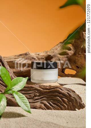A close-up of a glass cream jar mockup set on a piece of driftwood nestled in the sand, adorned with some fresh green leaves, on a warm orange backdrop. A minimalist image used to promote cosmetics. 121772070