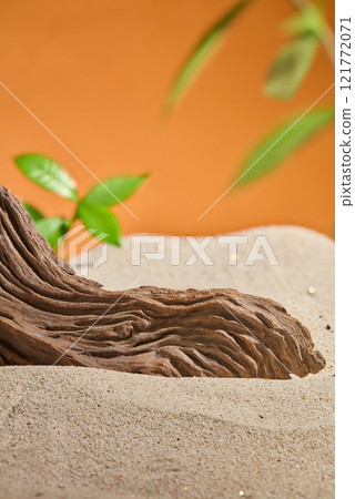 On the sand, a driftwood piece is set against an orange background with a few leaves scattered over it. A minimalist still life design used to showcase product ads. On the sand, a driftwood piece is set against an orange background with a few leaves scattered over it. A minimalist still life design used to showcase product ads. 121772071