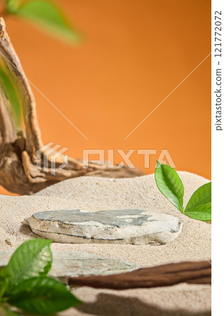 On the sand, an empty flat stone for displaying product is set against an orange background, with a driftwood piece and a few leaves scattered over it. A minimalist still life design for product ads. 121772072