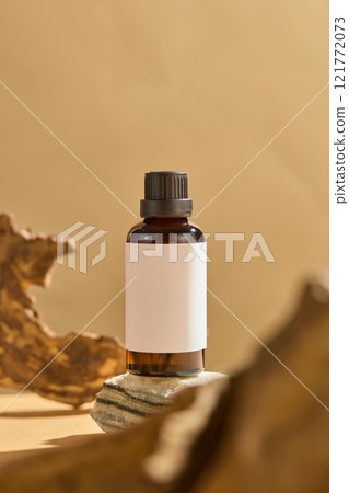 Frontal view of a dark-amber glass bottle with a black cap, sitting on a grey stone. A piece of wood surrounded the bottle, on a muted beige background. A minimalist picture for cosmetics advertising. Frontal view of a dark-amber glass bottle with a black cap, sitting on a grey stone. A piece of wood surrounded the bottle, on a muted beige background. A minimalist picture for cosmetics advertising. 121772073