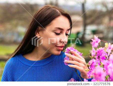 Russian woman in a blue sweater smelling pink rhododendron flowers in Primorsky Krai, Russia. The scene captures the essence of springtime and natural beauty Russian woman in a blue sweater smelling pink rhododendron flowers in Primorsky Krai, Russia. The scene captures the essence of springtime and natural beauty 121773576