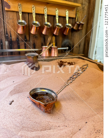 Copper cezve with fresh Turkish coffee on sandy stove, traditional copper coffee pots on wooden rack in background, symbolizing the authenticity of Middle Eastern coffee traditions. Copper cezve with fresh Turkish coffee on sandy stove, traditional copper coffee pots on wooden rack in background, symbolizing the authenticity of Middle Eastern coffee traditions. 121775451