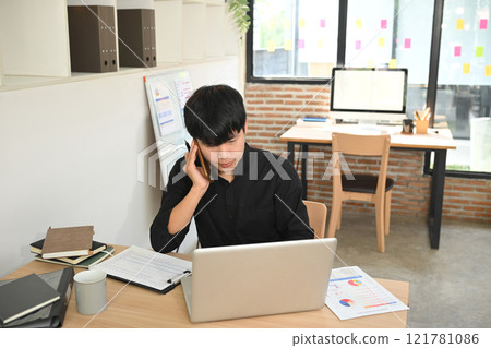Young businessman having pleasant phone conversation and using laptop at his office desk 121781086