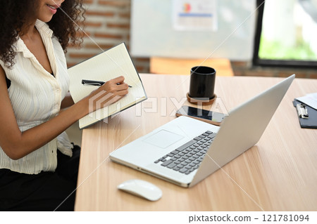 Young entrepreneur jotting down notes in a notebook while working on a laptop Young entrepreneur jotting down notes in a notebook while working on a laptop 121781094