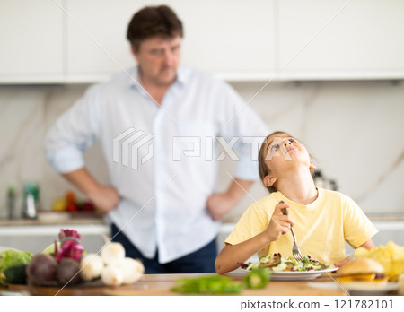 Father forcing little daughter to eat vegetable salad in kitchen 121782101