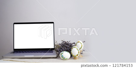 A laptop with a blank screen on a table festively decorated for the Easter holiday. Adorned working place with flowers and a basket with colorful eggs 121784153