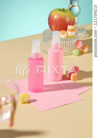 A top-down view of two mockups of pale pink spray bottles on a pale yellow surface; scattered around are colorful gummy candies, with a red apple on a clear glass pedestal. Pale teal background. A top-down view of two mockups of pale pink spray bottles on a pale yellow surface; scattered around are colorful gummy candies, with a red apple on a clear glass pedestal. Pale teal background. 121784215