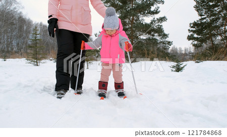 Young girl holds her ski poles with determination as her mother guides her through the snowy landscape during her first skiing experience 121784868