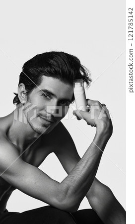 Handsome, smiling man sitting in casual pose and holding battle of cosmetic product for facial care against white background. Handsome, smiling man sitting in casual pose and holding battle of cosmetic product for facial care against white background. 121785142