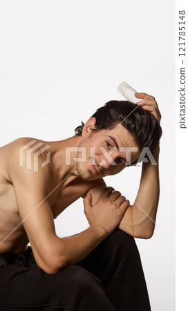 Stylish young man seated in relaxed pose, resting his head on his hand while holding bottle of cream, against white background. Stylish young man seated in relaxed pose, resting his head on his hand while holding bottle of cream, against white background. 121785148