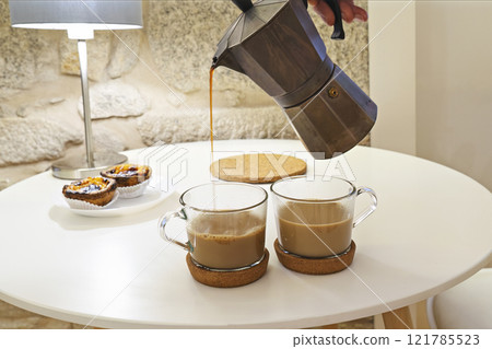 Moka pot pouring fresh coffee into glass mugs on cozy table with pastries, lamp, and stone wall background, creating warm ambiance Moka pot pouring fresh coffee into glass mugs on cozy table with pastries, lamp, and stone wall background, creating warm ambiance 121785523