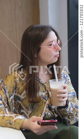 Pretty brunette with phone in hands relaxing in cafe. Woman drinking coffee cocktail Pretty brunette with phone in hands relaxing in cafe. Woman drinking coffee cocktail 121785832