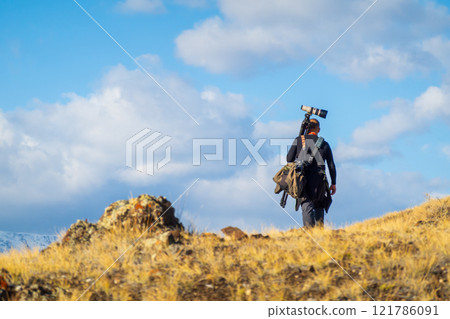 Photographer hiking with camera gear through grassy landscape under blue sky 121786091
