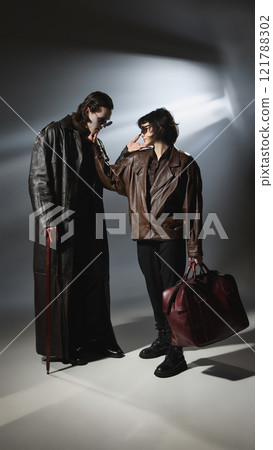 Vampire leans toward female companion in brown jacket, looks on her with love. Spotlight casts shadows on models in stylish outfits. Vampire leans toward female companion in brown jacket, looks on her with love. Spotlight casts shadows on models in stylish outfits. 121788302