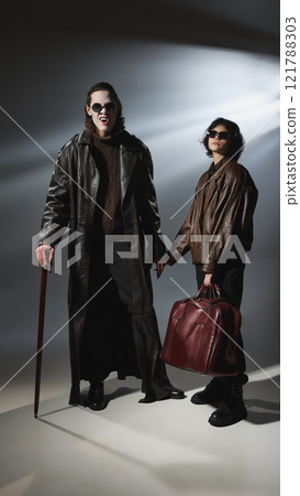Vampire laughing, showing sharp fangs, with female companion in brown leather jacket holding leather bag against lit background. 121788303