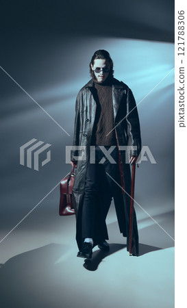 Vampire stands confidently in full leather attire holding cane in hand and bag. with a dim spotlight highlighting stylish outfit and stance. Vampire stands confidently in full leather attire holding cane in hand and bag. with a dim spotlight highlighting stylish outfit and stance. 121788306