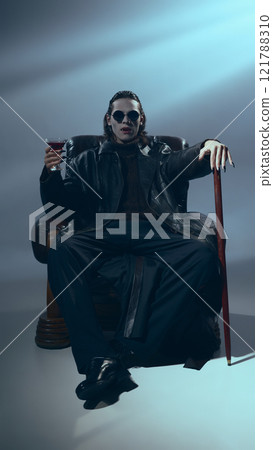 Man in leather jacket and sunglasses sits on chair, baring his sharp vampire fangs, holding drink glass and cane, illuminated by soft light. 121788310