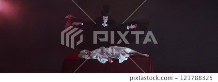 Banner. Vampire stands with arms spread wide, black cloak flowing, over table where woman dressed in white lies still, creating dramatic and eerie scene. 121788325
